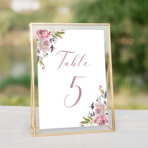 May include: A gold-framed table number card with a white background and pink floral design. The card reads "Table 5" in a cursive font.