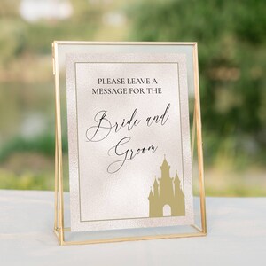 K&ouml;nnte beinhalten: Ein goldfarbener Rahmen mit einem wei&szlig;en Hintergrund und goldenem Glitzer. Das Schild sagt "Bitte hinterlassen Sie eine Nachricht f&uuml;r das Brautpaar" mit einer goldenen Silhouette eines Schlosses.