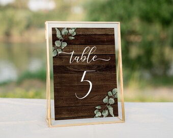 May include: A gold-framed table number sign with a rustic wood background and greenery accents. The sign reads "table 5".