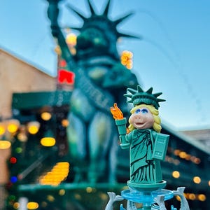 Puede incluir: Una figura de Lego verde de Miss Piggy vestida como la Estatua de la Libertad, sosteniendo una antorcha y de pie sobre una base de fuente con esculturas de peces. La figura est&aacute; delante de una Estatua de la Libertad m&aacute;s grande y desenfocada.