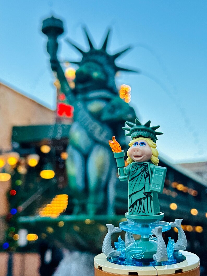 Puede incluir: Una figura de Lego verde de Miss Piggy vestida como la Estatua de la Libertad, sosteniendo una antorcha y de pie sobre una base de fuente con esculturas de peces. La figura est&aacute; delante de una Estatua de la Libertad m&aacute;s grande y desenfocada.