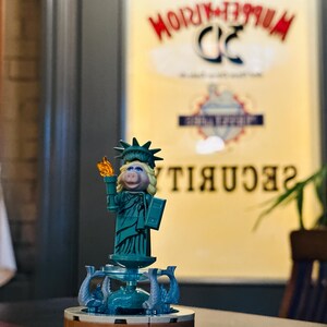 Puede incluir: Una figura de Lego verde y marr&oacute;n de Miss Piggy vestida como la Estatua de la Libertad. La figura sostiene una antorcha y un libro. La figura est&aacute; de pie sobre una base con una fuente y delfines. El fondo es una imagen borrosa de un cartel que dice "Muppetorium 3D Security".