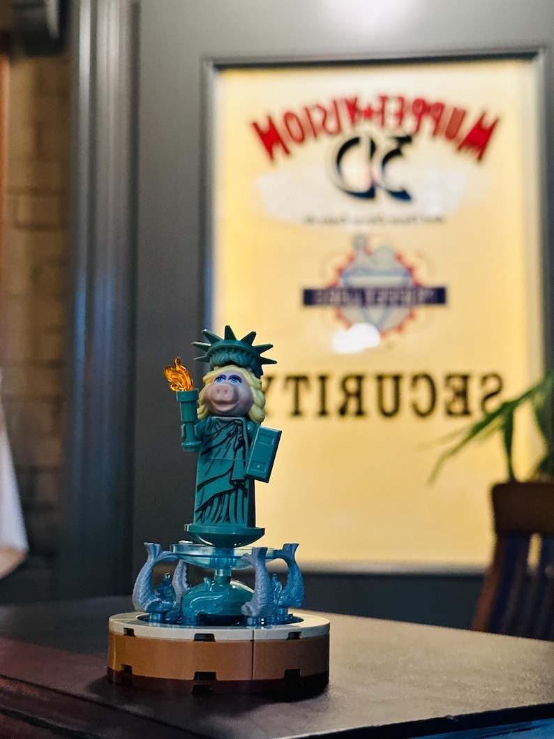 Puede incluir: Una figura de Lego verde y marr&oacute;n de Miss Piggy vestida como la Estatua de la Libertad. La figura sostiene una antorcha y un libro. La figura est&aacute; de pie sobre una base con una fuente y delfines. El fondo es una imagen borrosa de un cartel que dice "Muppetorium 3D Security".