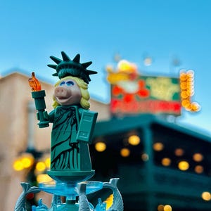 Puede incluir: Una figura de Lego verde de Miss Piggy vestida como la Estatua de la Libertad, sosteniendo una antorcha. La figura est&aacute; de pie sobre una base de fuente con agua azul y figuras de delfines grises.