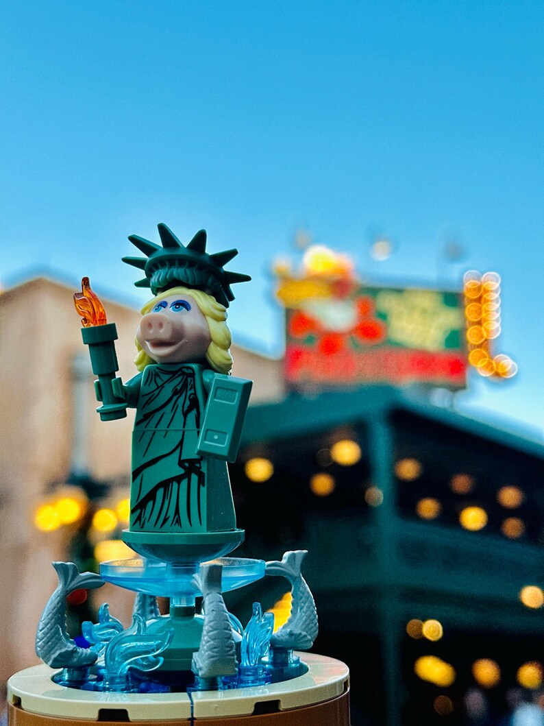 Puede incluir: Una figura de Lego verde de Miss Piggy vestida como la Estatua de la Libertad, sosteniendo una antorcha. La figura est&aacute; de pie sobre una base de fuente con agua azul y figuras de delfines grises.