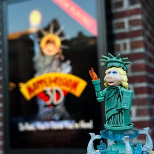 Puede incluir: Una figura de Lego verde y azul de Miss Piggy vestida como la Estatua de la Libertad. Ella sostiene una antorcha y est&aacute; de pie sobre una base con una fuente. El fondo es una pared de ladrillo con un cartel de pel&iacute;cula para Muppet Vision 3D.