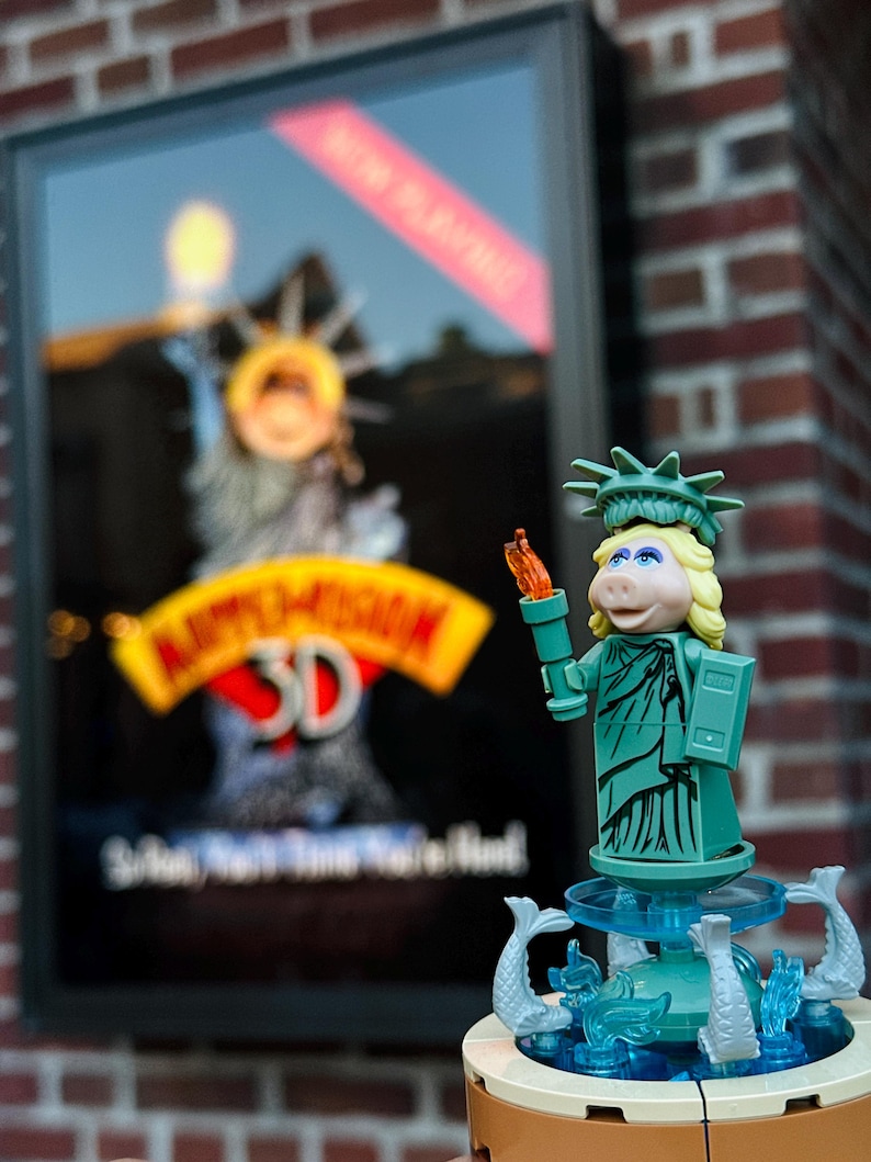 Puede incluir: Una figura de Lego verde y azul de Miss Piggy vestida como la Estatua de la Libertad. Ella sostiene una antorcha y est&aacute; de pie sobre una base con una fuente. El fondo es una pared de ladrillo con un cartel de pel&iacute;cula para Muppet Vision 3D.