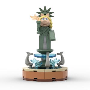 Puede incluir: Una figura de Lego verde de Miss Piggy vestida como la Estatua de la Libertad, de pie sobre una fuente con agua azul y figuras de delfines grises.