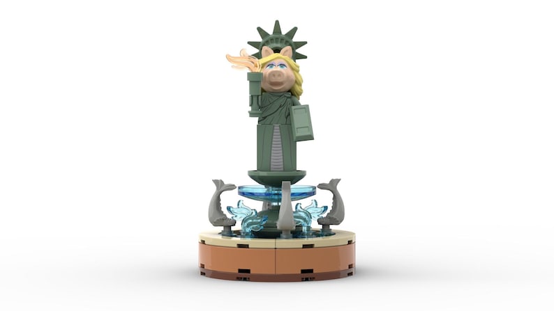 Puede incluir: Una figura de Lego verde de Miss Piggy vestida como la Estatua de la Libertad, de pie sobre una fuente con agua azul y figuras de delfines grises.