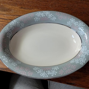 May include: An oval serving platter with a light gray border decorated with white and pink floral patterns. The center of the platter is a cream color, and the rim has a silver trim. This vintage dish is perfect for serving food.