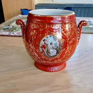 May include: A red and gold ceramic planter with a floral design and a scene of people in a garden. The planter has two handles and a wide base.
