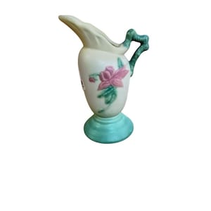 May include: A cream-colored ceramic pitcher featuring a painted pink flower with green leaves on the body. The pitcher has a textured green handle, a flared spout, and a solid green circular base. A small white sticker is visible on the side.