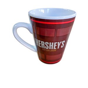 May include: A white-handled mug with a dark brown exterior featuring the Hershey's logo and the words "SPECIAL DARK MILDLY SWEET CHOCOLATE." The mug has a white interior and a red stripe near the rim. The mug's design resembles chocolate squares.
