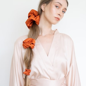 May include: A woman with long brown hair tied back with three orange scrunchies. She is wearing a light beige satin robe with a long sleeve and a sash tied at the waist.