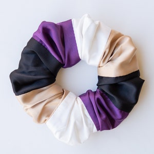 May include: A scrunchie with a circular design, featuring alternating stripes of black, tan, white, purple, and a darker purple. The fabric has a satin sheen, and the scrunchie is set against a white background. This hair accessory is designed for styling.