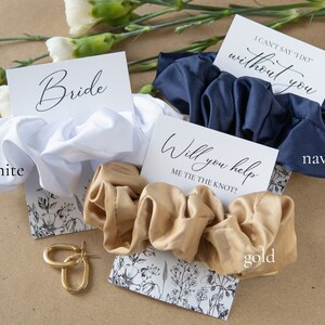 May include: Three satin scrunchies in white, navy blue, and gold with cards that say "Bride", "I can't say 'I do' without you", and "Will you help me tie the knot?"