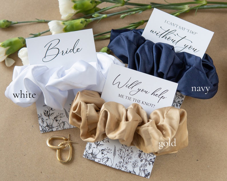 May include: Three satin scrunchies in white, navy blue, and gold with cards that say "Bride", "I can't say 'I do' without you", and "Will you help me tie the knot?"