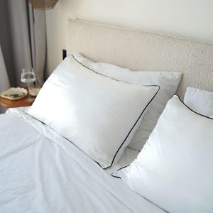 May include: A bed with white pillows featuring black edging. The pillows sit on a white sheet. The headboard is a neutral colour. The scene suggests a clean and minimalist bedroom setting.