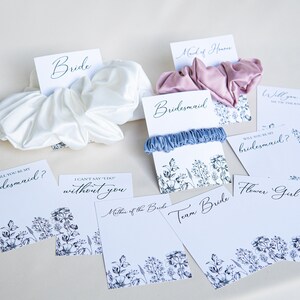 May include: A set of eight cards with floral designs and text, including "Bride", "Bridesmaid", "Maid of Honor", "Will you be my bridesmaid?", "I can't say 'I do' without you", "Will you help me tie the knot?", "Team Bride", and "Flower Girl".  Each card is paired with a satin scrunchie in a variety of colors.