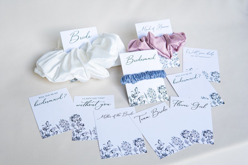 May include: A set of eight cards with floral designs and text, including "Bride", "Bridesmaid", "Maid of Honor", "Will you be my bridesmaid?", "I can't say 'I do' without you", "Will you help me tie the knot?", "Team Bride", and "Flower Girl".  Each card is paired with a satin scrunchie in a variety of colors.