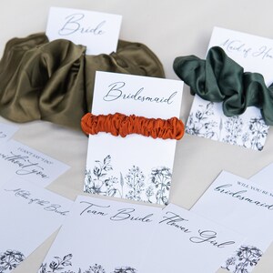 May include: A set of five cards with floral designs and the words "Bride", "Bridesmaid", "Maid of Honor", "Will you be my bridesmaid?" and "Will you help me tie the knot?"  The cards are paired with different colored scrunchies.