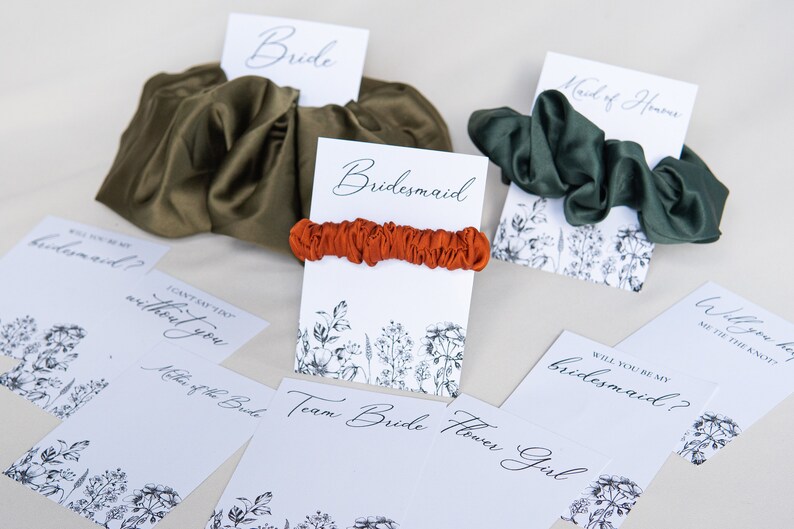 May include: A set of five cards with floral designs and the words "Bride", "Bridesmaid", "Maid of Honor", "Will you be my bridesmaid?" and "Will you help me tie the knot?"  The cards are paired with different colored scrunchies.