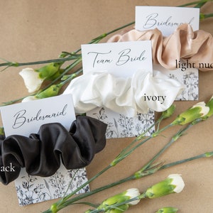 May include: Three satin scrunchies on cards with the words "Team Bride", "Bridesmaid", and "Bridesmaid". The scrunchies are in black, ivory, and light nude.
