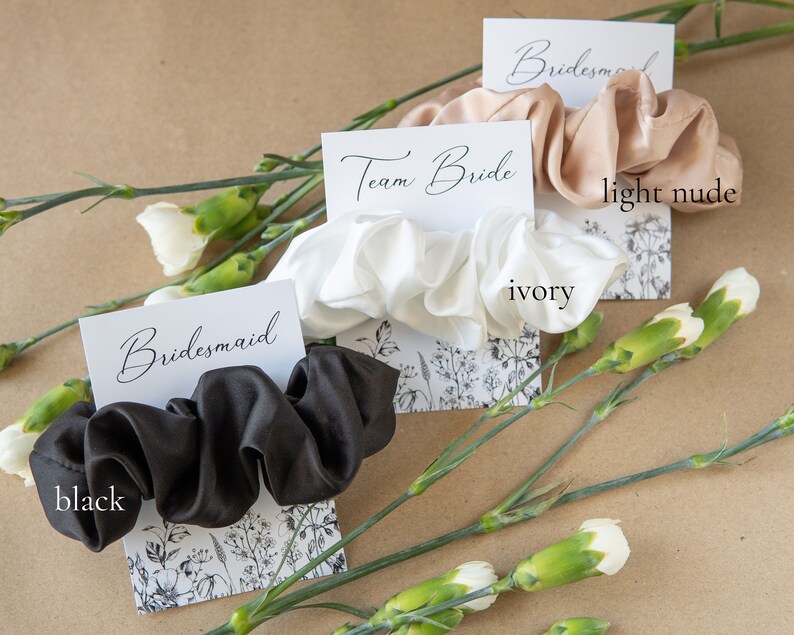 May include: Three satin scrunchies on cards with the words "Team Bride", "Bridesmaid", and "Bridesmaid". The scrunchies are in black, ivory, and light nude.
