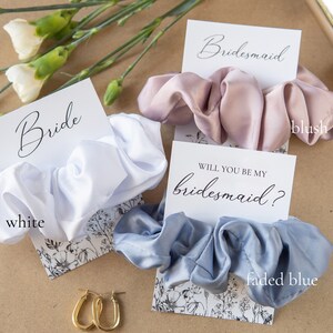 May include: Three satin scrunchies in white, blush pink, and faded blue, with cards that say "Bride", "Bridesmaid", and "Will you be my bridesmaid?"