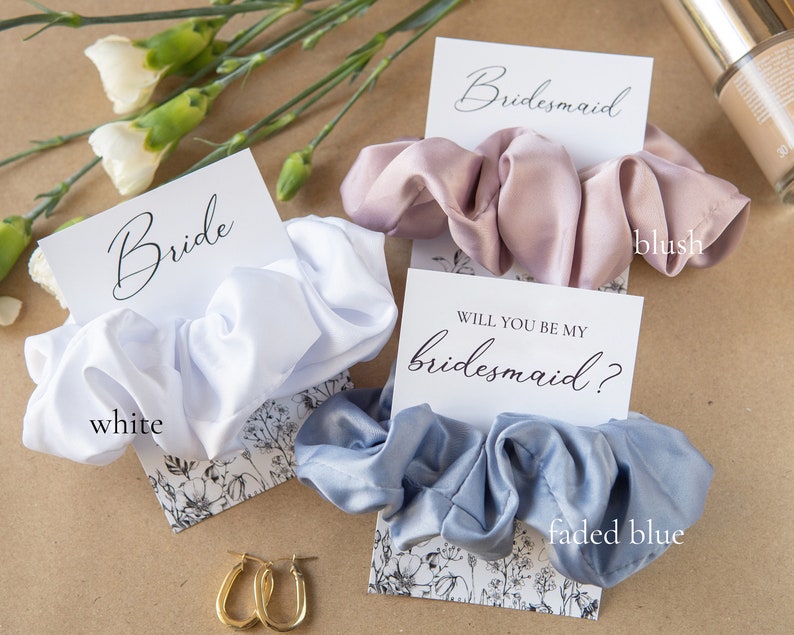 May include: Three satin scrunchies in white, blush pink, and faded blue, with cards that say "Bride", "Bridesmaid", and "Will you be my bridesmaid?"