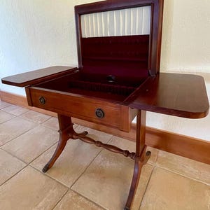 May include: A wooden antique table with a hinged top and a drawer. The table is made of dark wood and has a burgundy interior. The table has two side leaves and ornate legs.