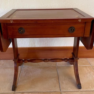 May include: A small, antique wooden side table with a rectangular top and drop leaves. The table has a single drawer with two round brass knobs. The legs are curved and connected by a stretcher. The table is stained a warm brown color.