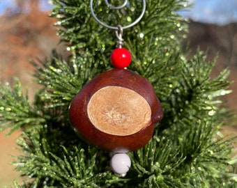 May include: A handmade Christmas ornament featuring a chestnut with a light brown center, accented by red and white beads. The ornament is suspended by a silver wire and set against a backdrop of a green Christmas tree.