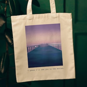 May include: A white canvas tote bag with a printed image of a pier extending into the ocean at sunset. The text "I guess I'll see you in the movies." is printed below the image.
