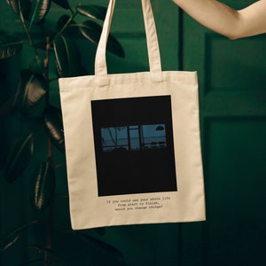 May include: A beige tote bag with black handles. The bag has a black square design with a window scene inside. The text on the bag reads: "If you could see your whole life from start to finish, would you change things?"