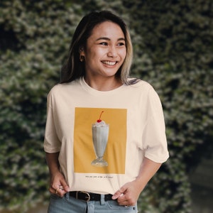 Könnte beinhalten: Ein weißes T-Shirt mit einem Aufdruck eines Milchshakes mit Schlagsahne und einer Kirsche obenauf. Der Aufdruck ist auf gelbem Hintergrund. Der Text unter dem Aufdruck lautet "Hast du gerade einen 5-€-Shake bestellt?"