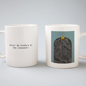 May include: Two white ceramic mugs with black text. One mug says "Sorry! My Prada's at the cleaners!" The other mug has a picture of a garment bag with a yellow tag that says "121".