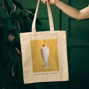 May include: A white canvas tote bag with a yellow background and a drawing of a milkshake with a cherry on top. The text on the bag reads "Did you just order a $5 shake?"