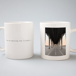 May include: Two white ceramic mugs. One mug has the text "You're waiting for a train..." printed on it. The other mug has a photo of a bridge with a road underneath it.