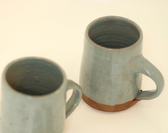 May include: Two ceramic mugs with a light blue glaze and a brown rim. The mugs are slightly different shapes and sizes.
