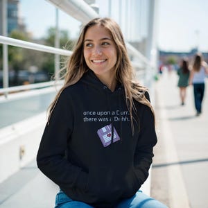 Peut inclure: Sweat à capuche noir avec le texte "once upon a Curr. there was a Duhh." et un graphique de disquette violette avec un cœur. Le sweat à capuche a une poche avant et une capuche à cordon.