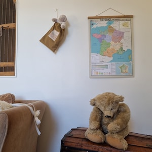 Może przedstawiać: Brązowy miś pluszowy siedzący na drewnianej skrzyni, za nim na ścianie wisi zabytkowa mapa Francji. Mapa nosi tytuł "La France - Les D&eacute;partements".