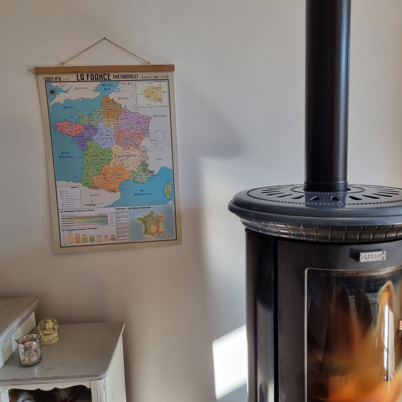 Może przedstawiać: Antyczna mapa Francji zatytułowana "La France Metropolitaine" wisi na białej ścianie obok czarnego kominka z przeszklonymi drzwiami. Mapa jest w odcieniach niebieskiego, zielonego i ż&oacute;łtego i przedstawia regiony Francji.