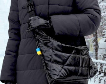Könnte beinhalten: Eine schwarze gesteppte Schultertasche mit glänzendem Finish. Die Tasche hat einen langen Riemen und wird über der Schulter einer Person getragen, die eine schwarze Steppjacke trägt.
