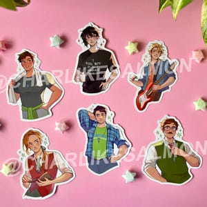 May include: A collection of die-cut stickers featuring illustrations of various characters. The stickers showcase individuals in diverse outfits and poses, with a pink background and star-shaped decorations. The text "POOL BOY AT THE VAMPIRE MANSION" is visible on one sticker.