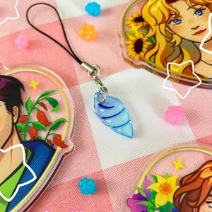 May include: A selection of clear acrylic charms and sweets on a pink and white checked surface. The charms display colourful illustrations of characters and flowers. A blue seashell charm hangs from a black lanyard. The Etsy shop name "charlikesalmon.etsy.com" is visible.