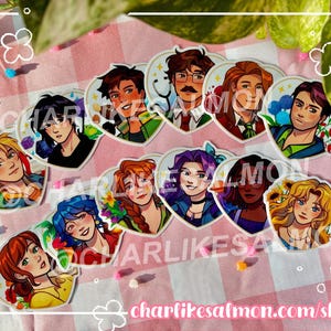 May include: A collection of heart-shaped stickers featuring colorful illustrations of various characters. The stickers showcase detailed artwork with vibrant colors and floral accents. The stickers are arranged on a pink and white checkered surface.