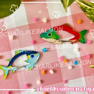 May include: Two fish-shaped carabiners with metal spring gates. One carabiner is red, green, and white, while the other is blue, yellow, and light blue. The text @CHARLIKESALMON and charlikesalmon.etsy.com is overlaid on the image.