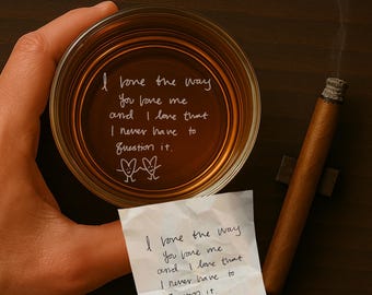 May include: A glass of amber liquid with a handwritten message on the bottom that reads "I love the way you love me and I love that I never have to question it."  Two stick figures with hearts are drawn below the message. A handwritten note with the same message is on a white piece of paper next to the glass. A lit cigar is on the table next to the glass.