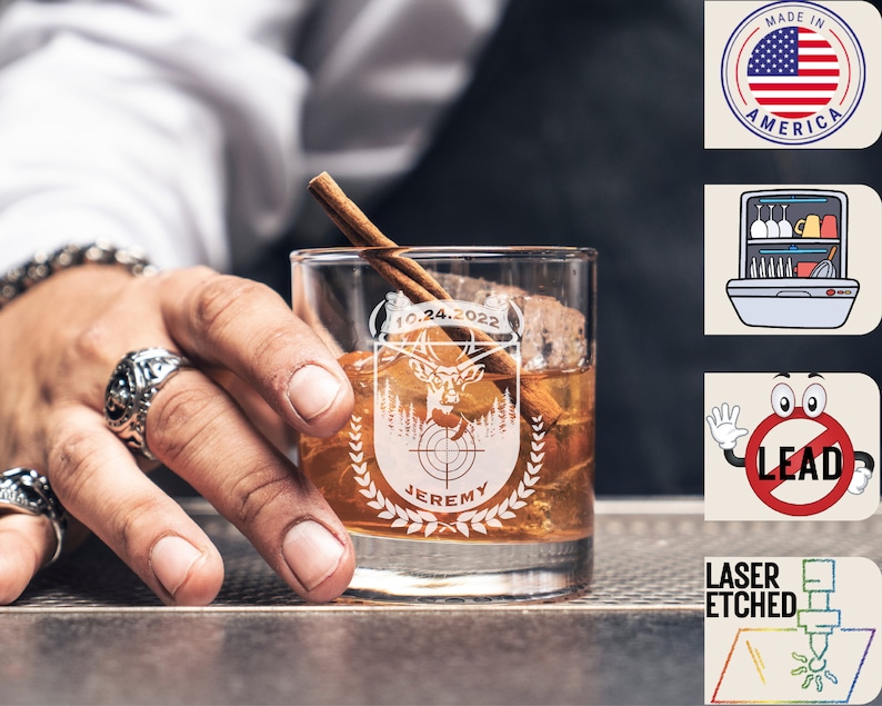 May include: A personalized whiskey glass with a laser etched design featuring a deer head, a target, and the text "10.24.2022" and "JEREMY". The glass is filled with a brown liquid and a cinnamon stick is resting on the rim.